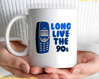 May include: White ceramic mug with a blue and white design. The mug features a blue cell phone with the text "90's Kid" on the screen and the text "Long Live The 90s" in blue letters.