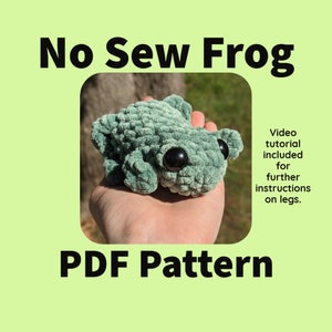 Op de afbeelding: Een groene gehaakte kikker met zwarte ogen. De kikker zit in de hand van een persoon. De afbeelding bevat de tekst "No Sew Frog PDF Pattern" en "Video tutorial included for further instructions on legs."