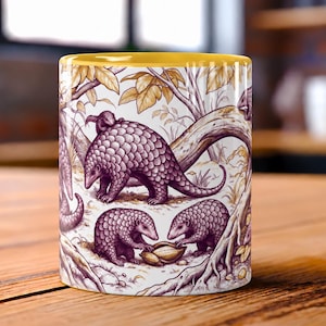 May include: A ceramic mug with a yellow interior and rim, featuring a detailed illustration of multiple pangolins in a forest setting. The artwork depicts the scaly creatures in shades of purple and brown among trees and foliage on a white background.