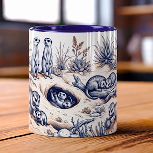 May include: A white ceramic mug with a deep blue interior rim featuring a detailed illustration of meerkats in a desert habitat. The design includes meerkats standing, resting in a burrow, and lounging on logs, surrounded by desert plants and rocks.