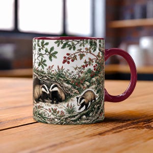 May include: A white ceramic mug with a deep burgundy handle and interior rim. The exterior features a detailed illustration of three badgers in a woodland setting with oak branches, green leaves, and clusters of red berries surrounding them.
