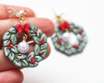 May include: Handmade Christmas wreath earrings. Each earring features a miniature wreath with green leaves, red berries, and a red bow. A dangling white pearl adds a touch of elegance. The earrings are attached to gold-colored hooks.
