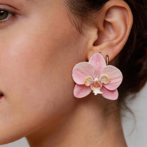 Op de afbeelding: Een paar roze orchidee bloem oorbellen. De oorbellen zijn lichtroze van kleur met een geel en roze centrum. De oorbellen zijn bevestigd aan een goudkleurige haak.