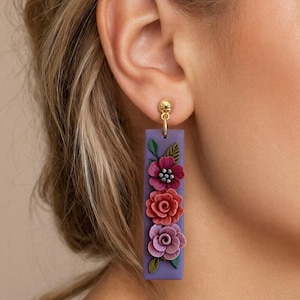 Op de afbeelding: Paar paarse rechthoekige oorbellen met driedimensionale bloemmotieven in rood, roze en paars. De oorbellen hebben een goudkleurige oorbel. De bloemen zijn gedetailleerd en getextureerd.