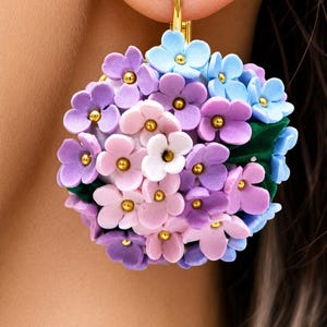 Op de afbeelding: Een ronde bloemenoorbel met een trosje kleine, gebeeldhouwde bloemen in tinten lichtblauw, lavendel, roze en wit. Elke bloem heeft een klein goudkleurig hartje. Het geheel bevat kleine donkergroene blaadjes en hangt aan een goudkleurige haak.