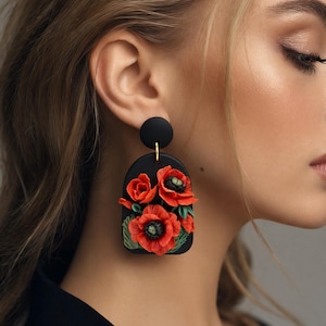 Op de afbeelding: Zwarte, bloemvormige oorbellen met felrode klaprozen en groene bladeren. De oorbellen hebben een zwart boogontwerp met een goudkleurige pin en een cirkelvormige bovenkant. Een uniek accessoire.
