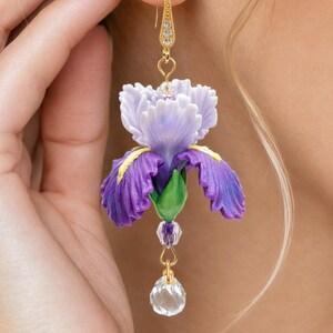 Op de afbeelding: Een hangende oorbel met een gebeeldhouwde paarse en lichtlavendelkleurige irisbloem met groene bladeren en gele accenten. De bloem hangt aan een goudkleurige haak met kleine heldere kristallen en een heldere gefacetteerde kristallen druppel.