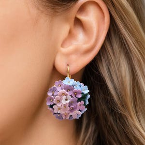 Op de afbeelding: Een close-up van een bloemenoorbel. De oorbel heeft een cluster van kleine, delicate bloemen in tinten paars, roze en blauw. De bloemen zijn gerangschikt in een bolvorm en bevestigd aan een goudkleurige ring.