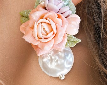 May include: A floral earring featuring a large peach-colored rose, a smaller lavender rose, and green leaves. A large, white, pearl-shaped bead is below the flowers. A small silver bead is at the bottom of the pearl.