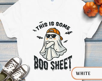 May include: White t-shirt with a Halloween-themed graphic. The design features a cartoon ghost wearing sunglasses and a baseball cap, with the text "THIS IS SOME BOO SHEET" in black. Bats are also included in the design.