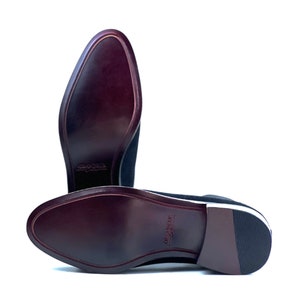 Peut inclure: Une paire de chaussures en daim bleu marine fonc&eacute; avec des semelles lisses et polies d'un bordeaux profond. Les semelles portent le mot "GRANDEUR" en relief au centre. Les talons sont empil&eacute;s avec une finition marron fonc&eacute; et un patin.