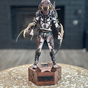 May include: A detailed action figure of a Predator with a brown and white color scheme. The figure is standing on a hexagonal base with the word "PREDATOR" printed on it.