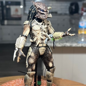 May include: A detailed action figure of a Predator, a fictional alien hunter. The figure is wearing a camouflage suit with a detailed mask and weapons. The figure is posed in a dynamic stance, with one arm raised and the other holding a weapon.