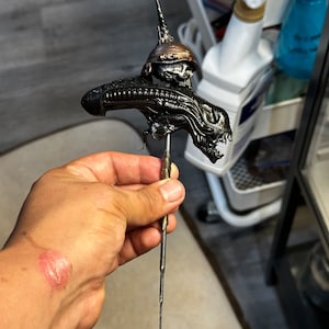 May include: A detailed, metallic sculpture of an alien creature, featuring a long, spiked head and a menacing, open-mouthed face. The sculpture is held by a hand, showcasing its intricate design and dark, bronze color.