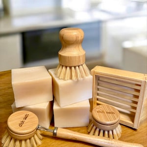 Può includere: Una collezione di articoli per lavare i piatti ecologici. L'immagine presenta diverse saponette rettangolari, due spazzole per piatti con manici in legno e un portasapone in legno. Sulle spazzole e sul portasapone è stampata la scritta "dish suds".