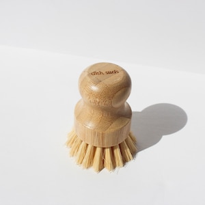 Puede incluir: Un cepillo para platos de madera con cerdas naturales. El cepillo tiene un mango de bambú con las palabras "dish suds" grabadas en la parte superior.