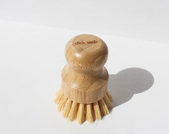 May include: A wooden dish brush with natural bristles. The brush has a bamboo handle with the words "dish suds" engraved on the top.