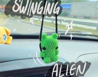 Puede incluir: Una muñeca alienígena verde tejida a ganchillo cuelga de una cuerda negra, con la palabra "SWINGING" encima y "ALIEN" debajo. La muñeca tiene grandes ojos negros y está delante del salpicadero de un coche.