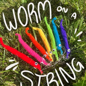 May include: A collection of colorful, crocheted worms on a string, arranged on a bed of green grass. The worms are in various bright colors, including red, pink, orange, yellow, green, blue, and purple. The words "WORM on a STRING" are written in white.