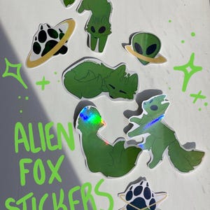 May include: A collection of green Alien Fox stickers with various designs, including alien heads, foxes, and planets. The stickers have a holographic or glossy finish. The text "ALIEN FOX STICKERS" is written in green.