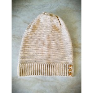 Puede incluir: Un gorro de punto beige con textura de canal&eacute; y tres botones naranjas en el lateral.