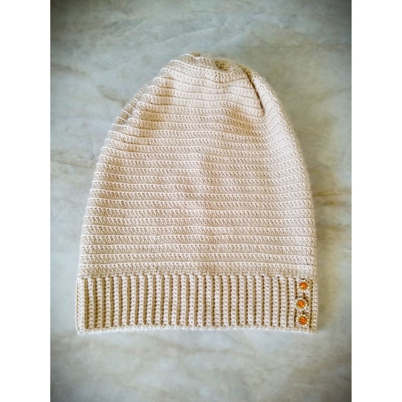 Puede incluir: Un gorro de punto beige con textura de canal&eacute; y tres botones naranjas en el lateral.