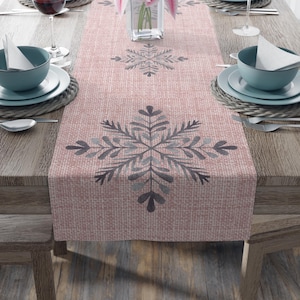 May include: A pink table runner with a snowflake pattern. The runner is made of a textured material and features large grey snowflakes. The table is set with teal bowls, plates, and wine glasses, creating a festive dining scene.