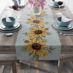 Può includere: Una tovaglia azzurra con un motivo a girasoli. La tovaglia presenta grandi girasoli gialli con centri marroni, foglie verdi e fiori gialli più piccoli. La tavola è apparecchiata con ciotole, piatti, calici e posate turchesi.