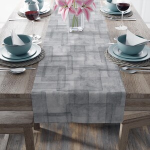 May include: A gray and white geometric patterned table runner with a subtle watercolor effect. The runner is laid out on a wooden table with place settings on either side.