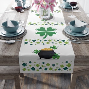 May include: A white table runner featuring a green shamrock design and a pot of gold. The runner is decorated with green shamrocks of various sizes and a pot of gold filled with gold coins.