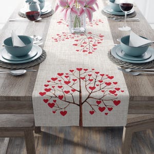May include: A beige table runner with a red heart tree design. The runner is decorated with two smaller heart trees at the top and a larger heart tree in the center. The runner is on a wooden table with two place settings on each side.