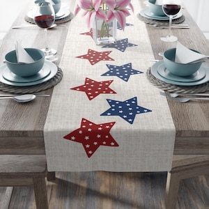 May include: A white linen table runner with red and blue stars. The stars are filled with white polka dots. The table runner is on a wooden table with two place settings.