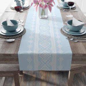 May include: A light blue table runner with a white geometric pattern. The runner is placed on a wooden table set for a meal, with plates, bowls, silverware, and wine glasses. A vase of pink flowers sits in the center.