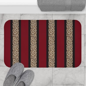 May include: A rectangular bath mat with a bold design. It features alternating vertical stripes of burgundy, black, and leopard print. The leopard print has a tan background with brown spots. The mat is on a tiled floor.