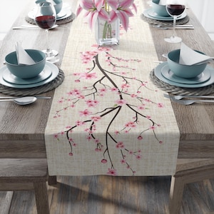 Könnte beinhalten: Beiger Tischläufer mit einem floralen Design aus rosa Kirschblüten auf dunkelbraunen Ästen. Der Läufer liegt auf einem Holztisch mit Gedecken und einer Vase mit rosa Lilien.