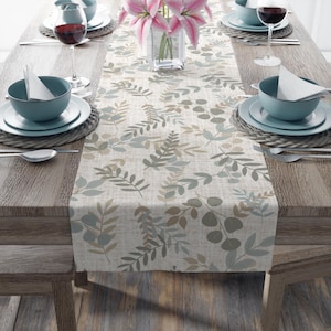 May include: A beige table runner with a leafy pattern in shades of green and brown. The runner is laid on a wooden table with two place settings on each side.