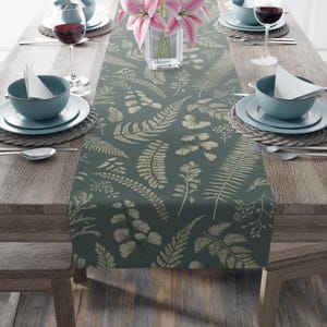 May include: A dark teal table runner with a botanical fern and leaf pattern in beige. The runner is placed on a wooden table set for a meal, with plates, bowls, silverware, and wine glasses. A vase of pink flowers is in the center.