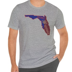 May include: A grey t-shirt with a graphic of the state of Florida in blue and maroon, with the words "HOME" and "DIVIDED" and a football in the centre. The shirt has short sleeves.