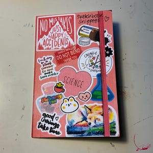 以下が含まれることがあります： ピンク色のスケッチブック。ステッカーには「No Mistakes Just Happy Accidents」、「Each day is a blank canvas」、そして「Science」という言葉が書かれています。その他、眠っているキツネ、クマ、ビーカーのステッカーも付いています。