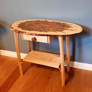 May include: Oval wooden side table with a natural edge top and a small drawer. The table has a lower shelf and is supported by four legs. The top is approximately 30 inches long.