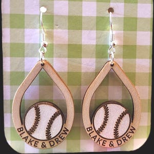 May include: A pair of wooden baseball earrings. Each earring features a teardrop-shaped frame with a baseball design in the center. The baseballs are white with brown stitching, and the text "BLAKE & DREW" is printed around the baseballs. Silver-colored ear hooks.