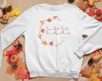 Puede incluir: Sudadera blanca con la frase "I've Got Fall On My Mind" en cursiva. El diseño incluye una corona de hojas de otoño en tonos naranja y rojo. La imagen está adornada con decoraciones otoñales.
