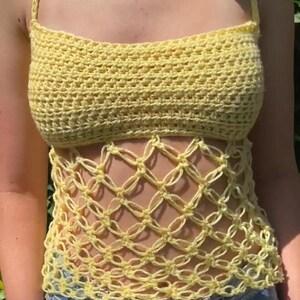 May include: A handmade, yellow crochet top with a halter neck and open weave design. The top is a light yellow color and is worn with blue denim jeans. The crochet work is visible and detailed, showcasing the craftsmanship of the garment.