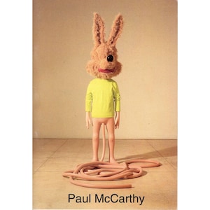 May include: A whimsical figure with a rabbit head, wearing a yellow long-sleeve shirt and pink shorts. The figure's legs extend into a series of pink, tube-like appendages. The text "Paul McCarthy" is at the bottom.