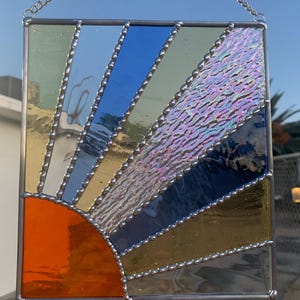 May include: A stained glass sunburst window hanging with a silver chain. The sunburst is made of orange, yellow, blue, and green glass with a textured iridescent center. The glass pieces are outlined with silver beads.