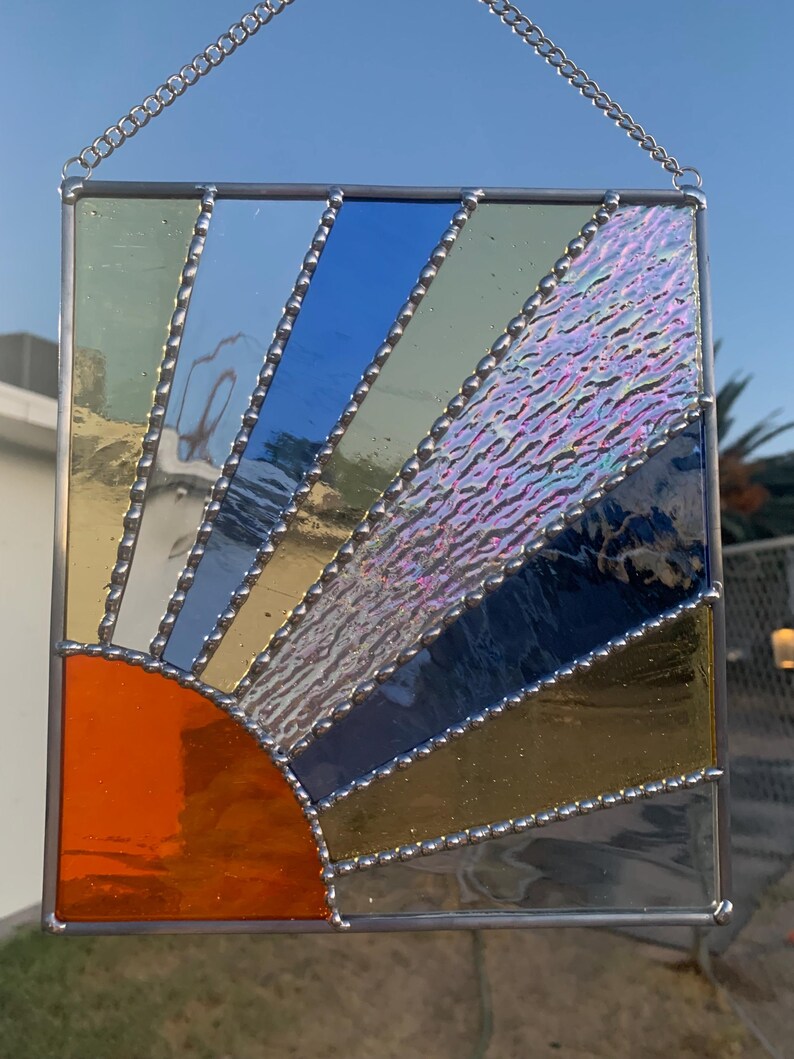 May include: A stained glass sunburst window hanging with a silver chain. The sunburst is made of orange, yellow, blue, and green glass with a textured iridescent center. The glass pieces are outlined with silver beads.