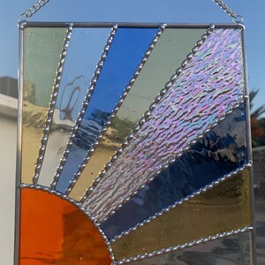 May include: A stained glass sunburst window hanging with a silver chain. The sunburst is made of iridescent glass in shades of blue, green, and orange.