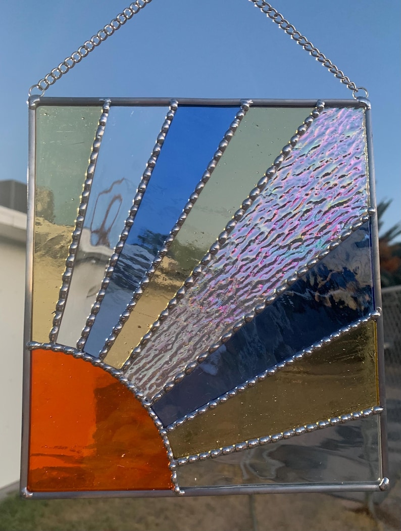 May include: A stained glass sunburst window hanging with a silver chain. The sunburst is made of iridescent glass in shades of blue, green, and orange.