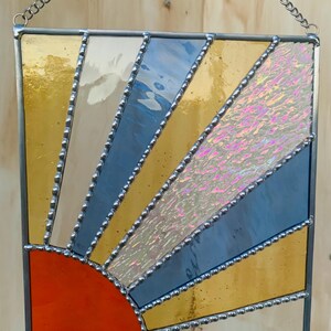 May include: A stained glass sunburst with a red, orange, yellow, blue, and iridescent white design. The sunburst is framed in silver and has a chain for hanging.