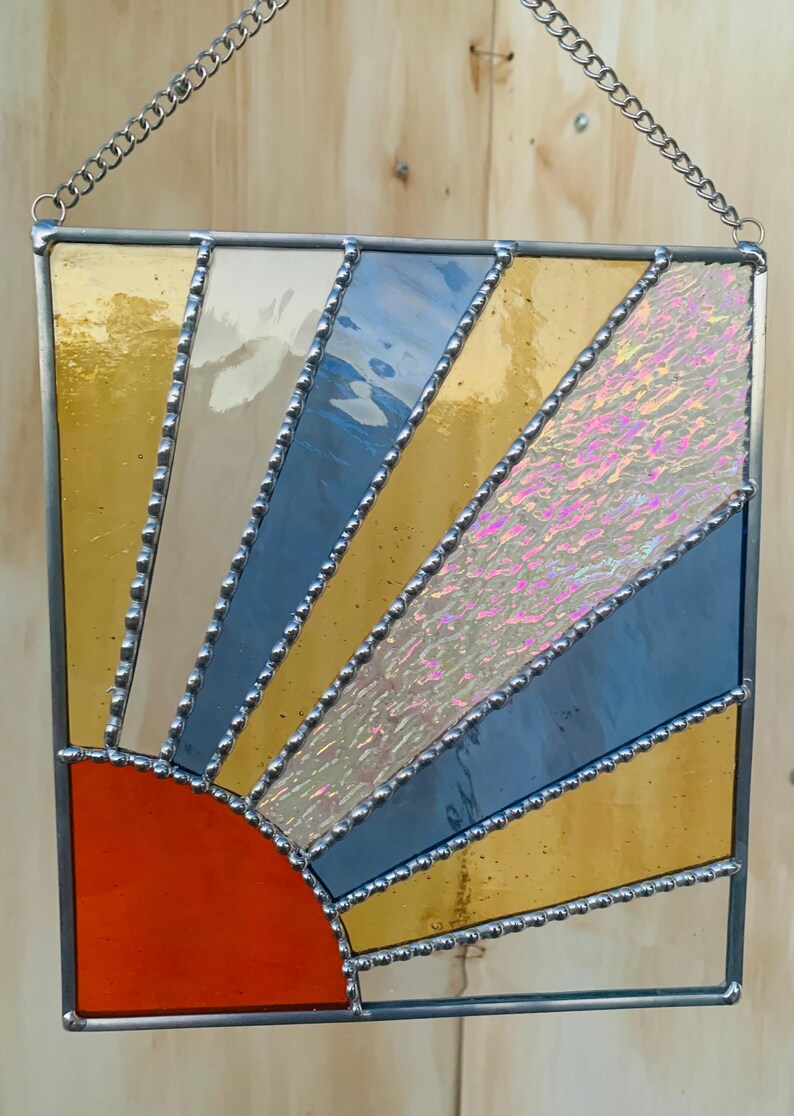May include: A stained glass sunburst with a red, orange, yellow, blue, and iridescent white design. The sunburst is framed in silver and has a chain for hanging.